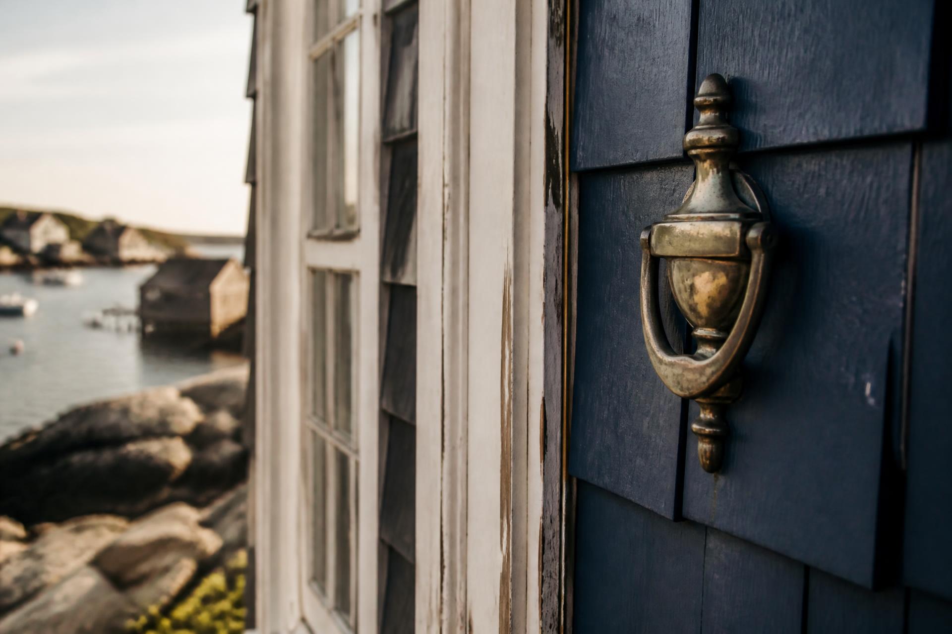 A weathered brass door knocker on a deep navy shingled coastal home, with a rocky New England harbor beyond.